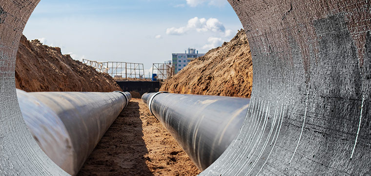 Construction site with two large metal pipes in a trench, viewed from inside another pipe.