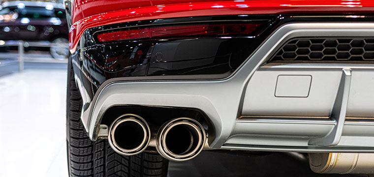 A close-up view of the rear end of a red car, highlighting its dual exhaust pipes and part of the bumper. The exhaust pipes are metallic and cylindrical, positioned side by side. The bumper features black and silver elements with a honeycomb pattern on one section. The car is parked indoors, with another vehicle partially visible in the background.