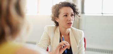 Image shows a women wearing business attire holding a pen. She appears in mid conversation with multiple other people.