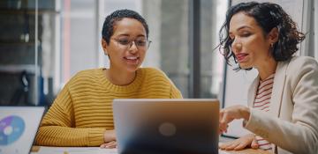 Image shows two women in front of a laptop. Working together, one referring to the laptop in front whilst the other professional listens