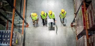 Image shows an overhead shot in a warehouse. There are four people in the centre of an isle of racking. They are wearing high visibility vests and yellow hardhats.
