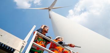 Image shows a wind turbine from standing beneath it. With two men in orange high visability jackets who look at be working  on the wind turbine.