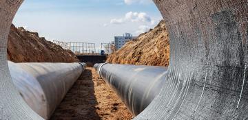Image shows construction site with two large metal pipes in a trench, viewed from inside another pipe.