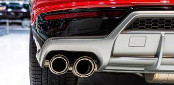 A close-up view of the rear end of a red car, highlighting its dual exhaust pipes and part of the bumper. The exhaust pipes are metallic and cylindrical, positioned side by side. The bumper features black and silver elements with a honeycomb pattern on one section. The car is parked indoors, with another vehicle partially visible in the background.