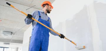 A construction worker in a blue overall and orange hard hat uses a long-handled paint roller to apply white paint on an interior wall of an unfinished building with exposed concrete ceilings and visible wiring. The worker's face is blurred for privacy.